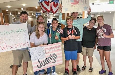 Happy group of 8 people at the airport, with balloons, signs, and small USA flags, all to welcome their international student.