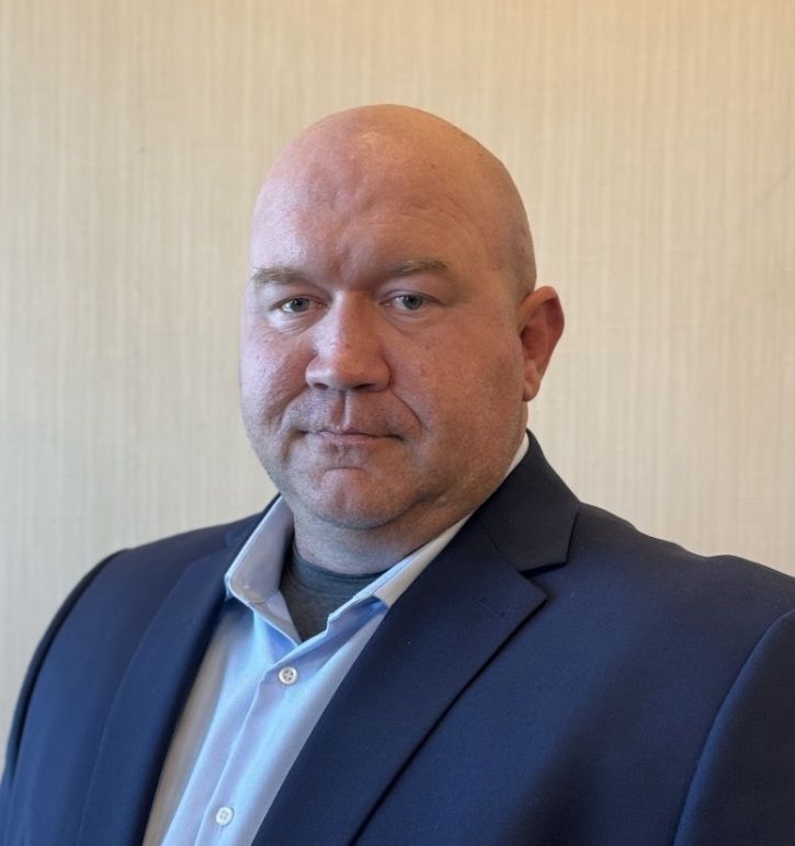 A bald man in a suit and blue shirt is standing in front of a white wall.