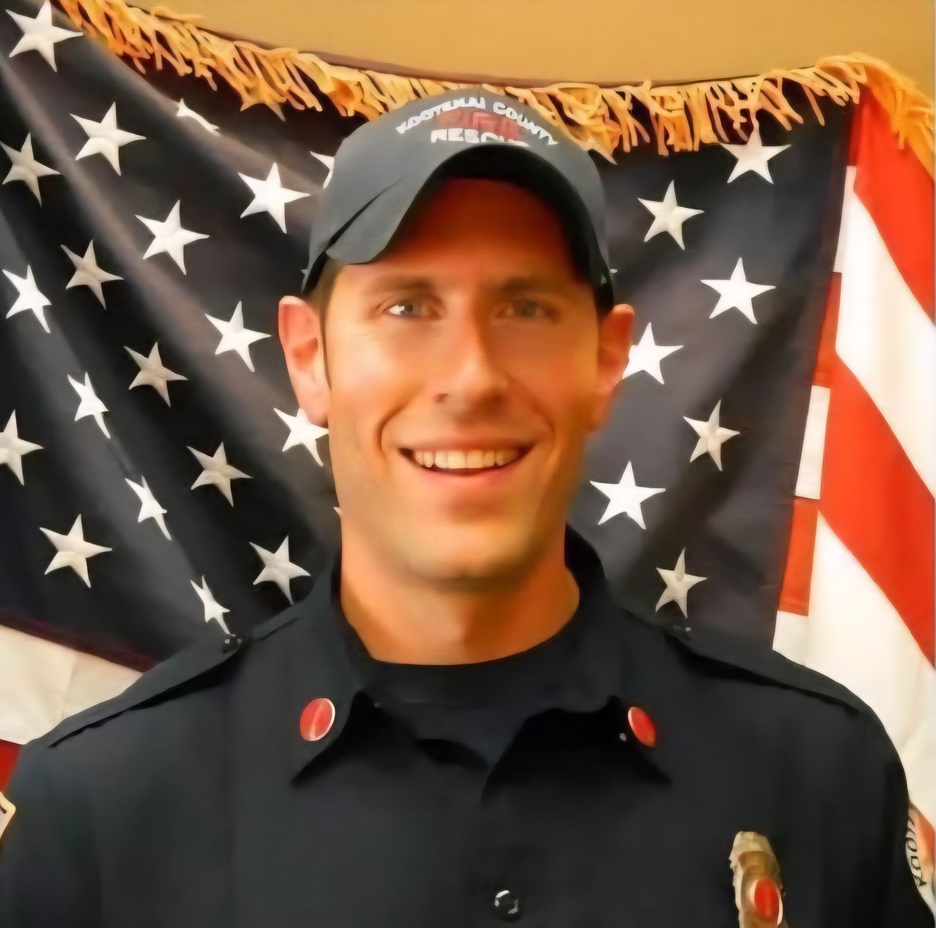Firefighter in uniform smiling, in front of an American flag. He wears a cap and has red lapel pins.