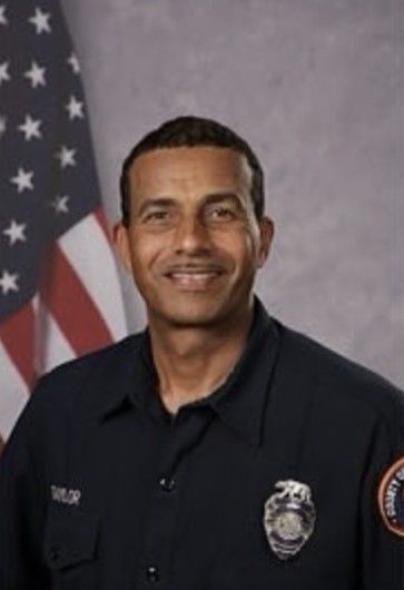 A man in a police uniform is smiling in front of an american flag. James O. Taylor