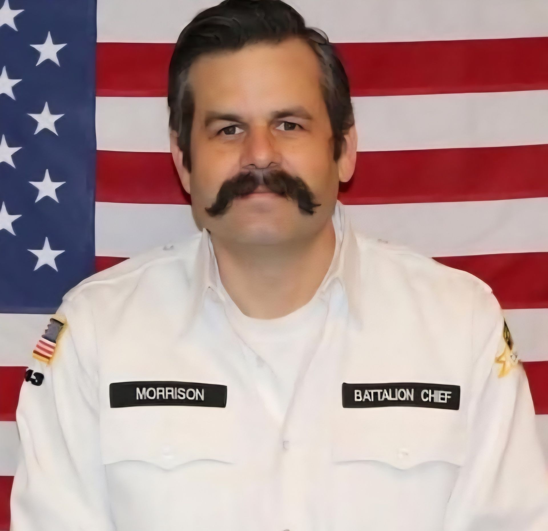Battalion Chief Morrison in uniform with a large mustache, American flag background.