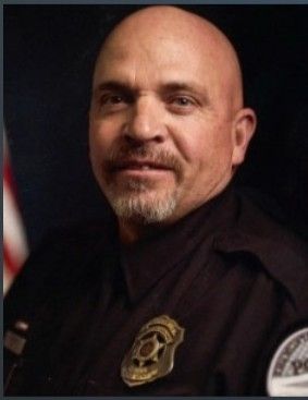 Bald, older man in a police uniform with a badge, smiling slightly.