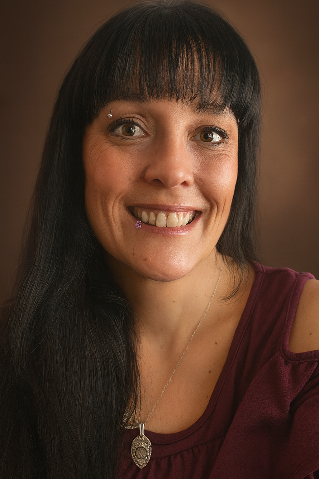 Woman with long dark hair, bangs, smiling, wearing a maroon top, with eyebrow and lip piercings.