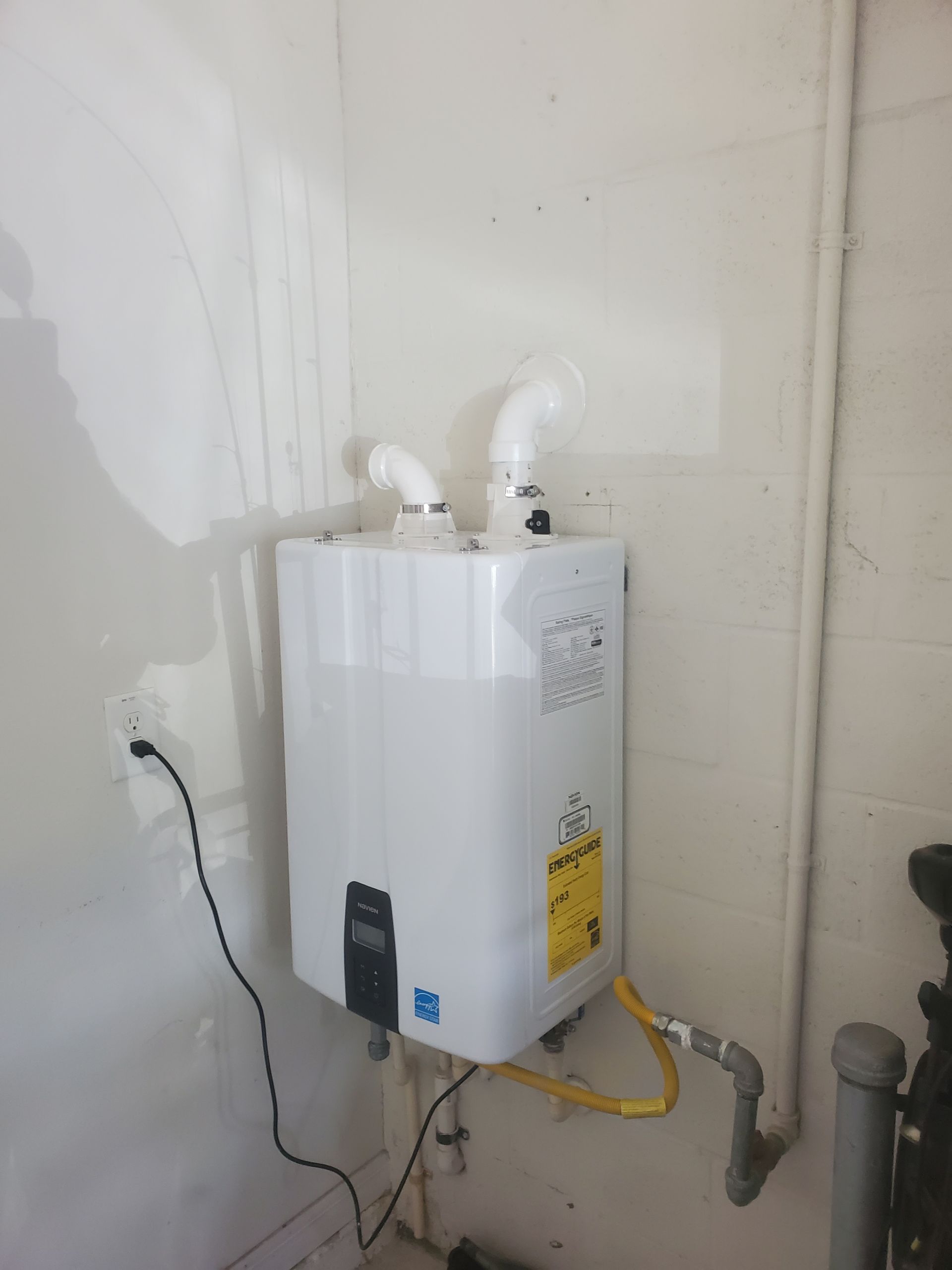 White tankless water heater on a white wall; white exhaust pipes; yellow gas line; black control panel.