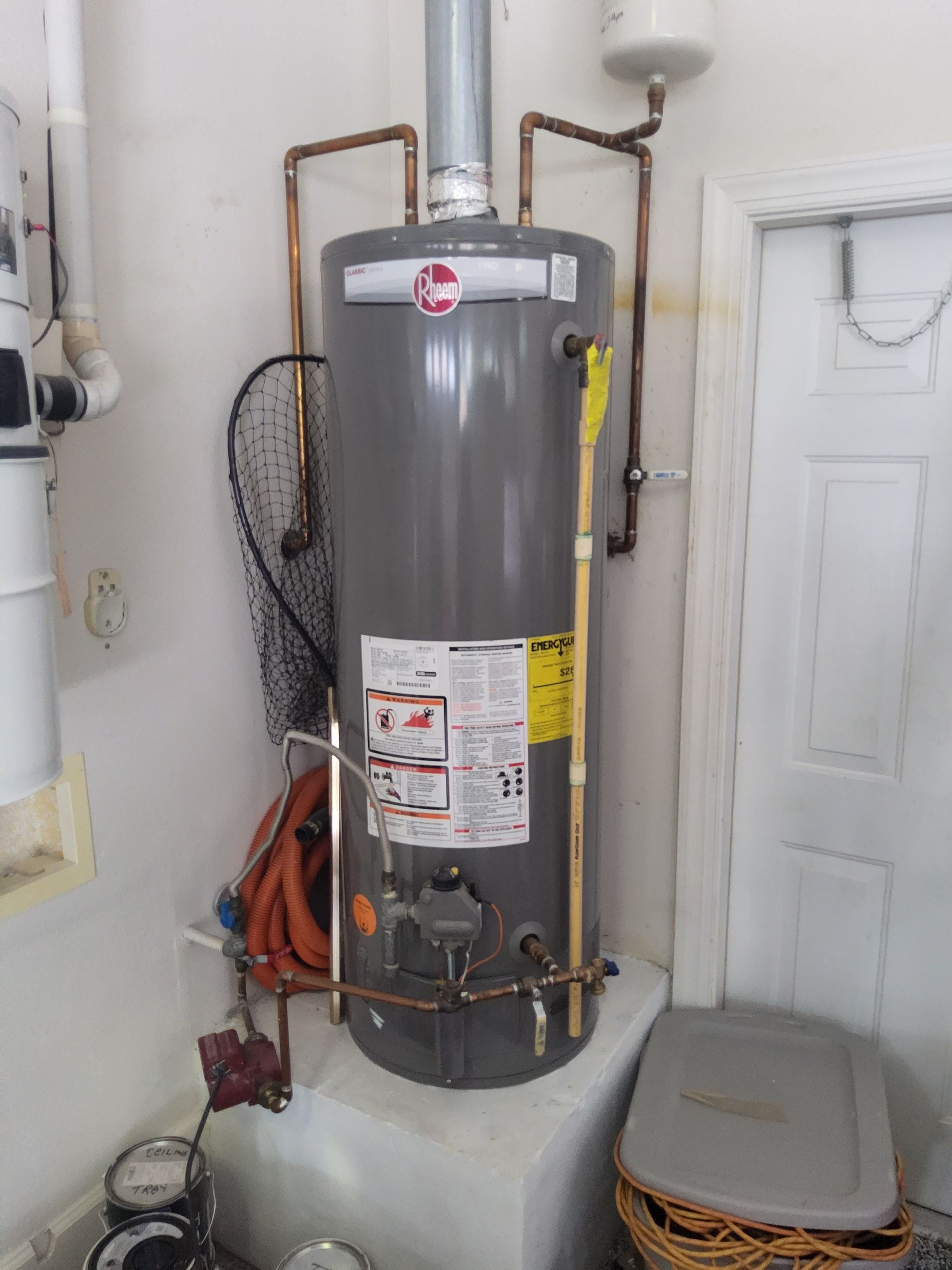 A tall, gray water heater in a utility room, with copper pipes and a white door in the background.