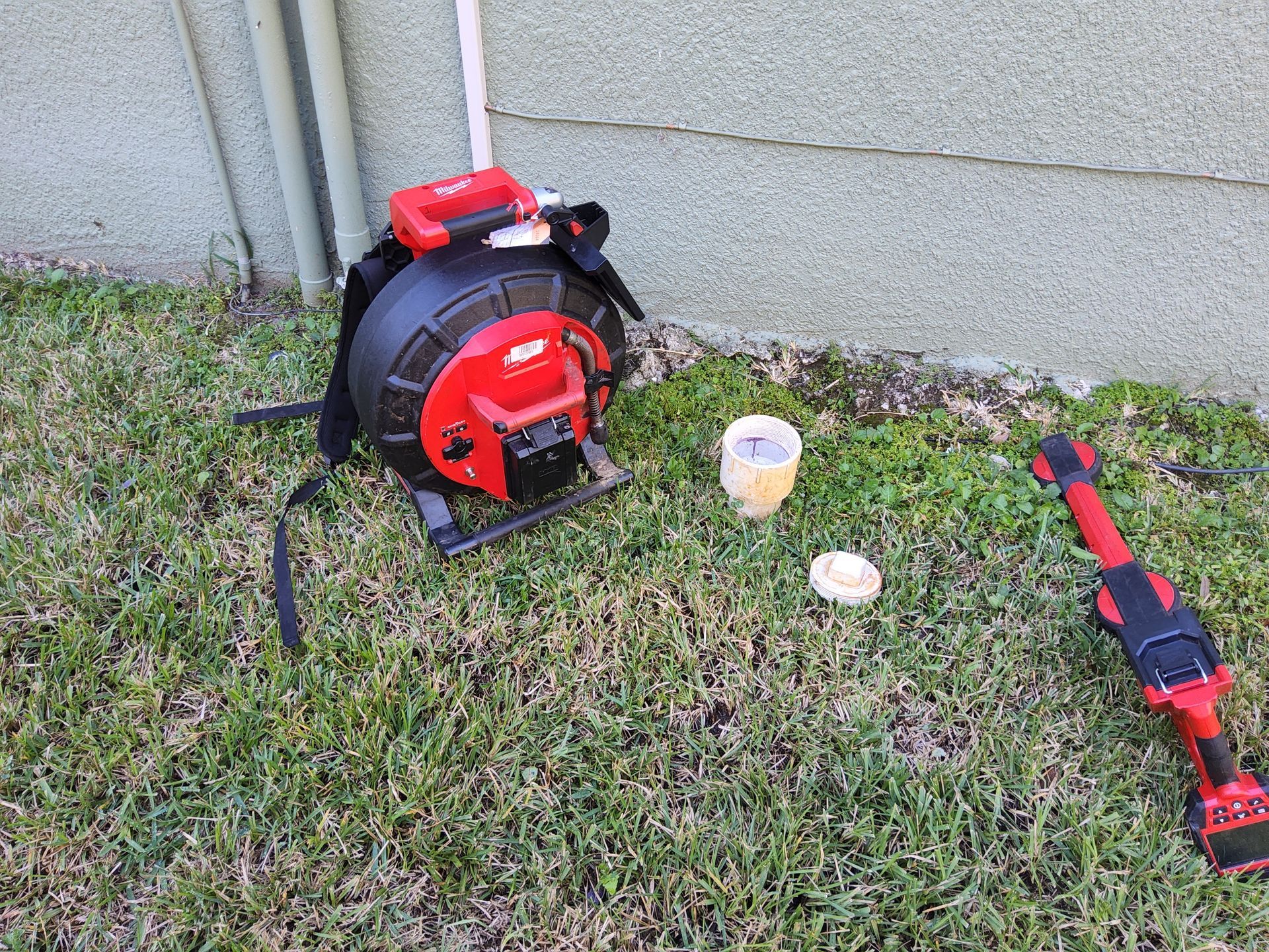 A red and black cable reel with a backpack strap and a red and black tool on grass near a building.