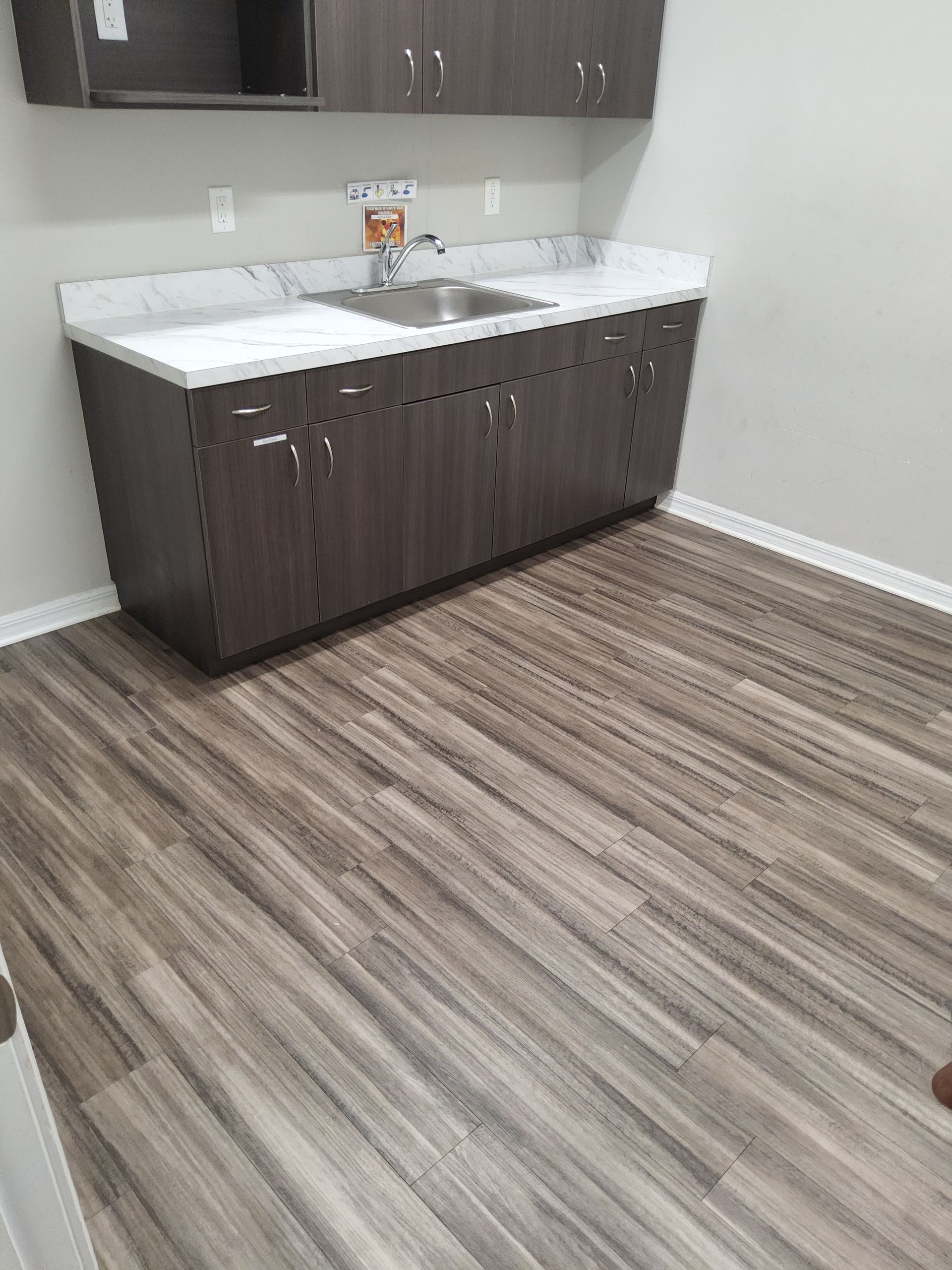 A room with gray cabinets, sink, and wood-look flooring. White countertop. Wall outlets.
