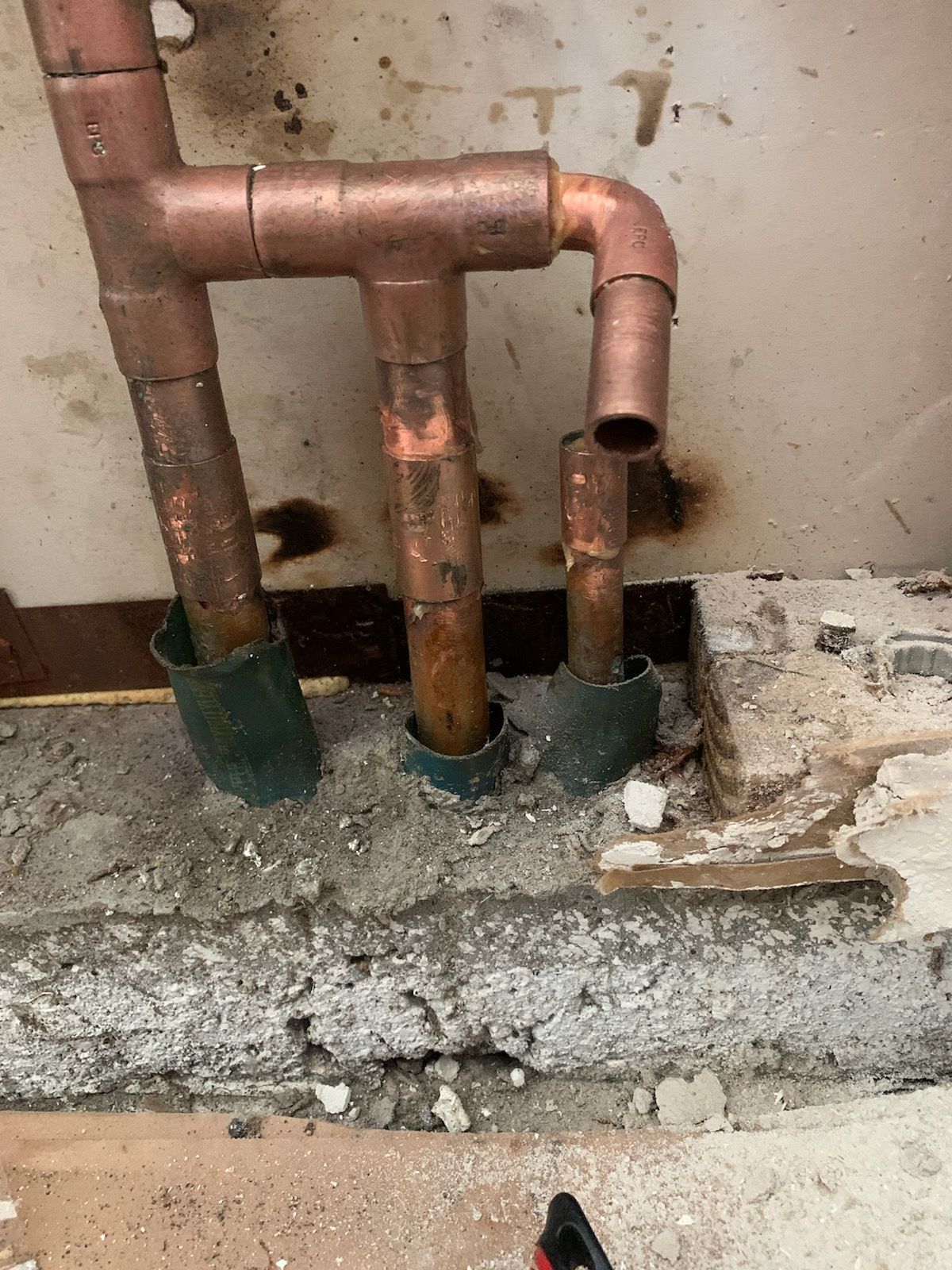 Copper pipes rising from a concrete floor, with a tee fitting and elbow, showing signs of wear.