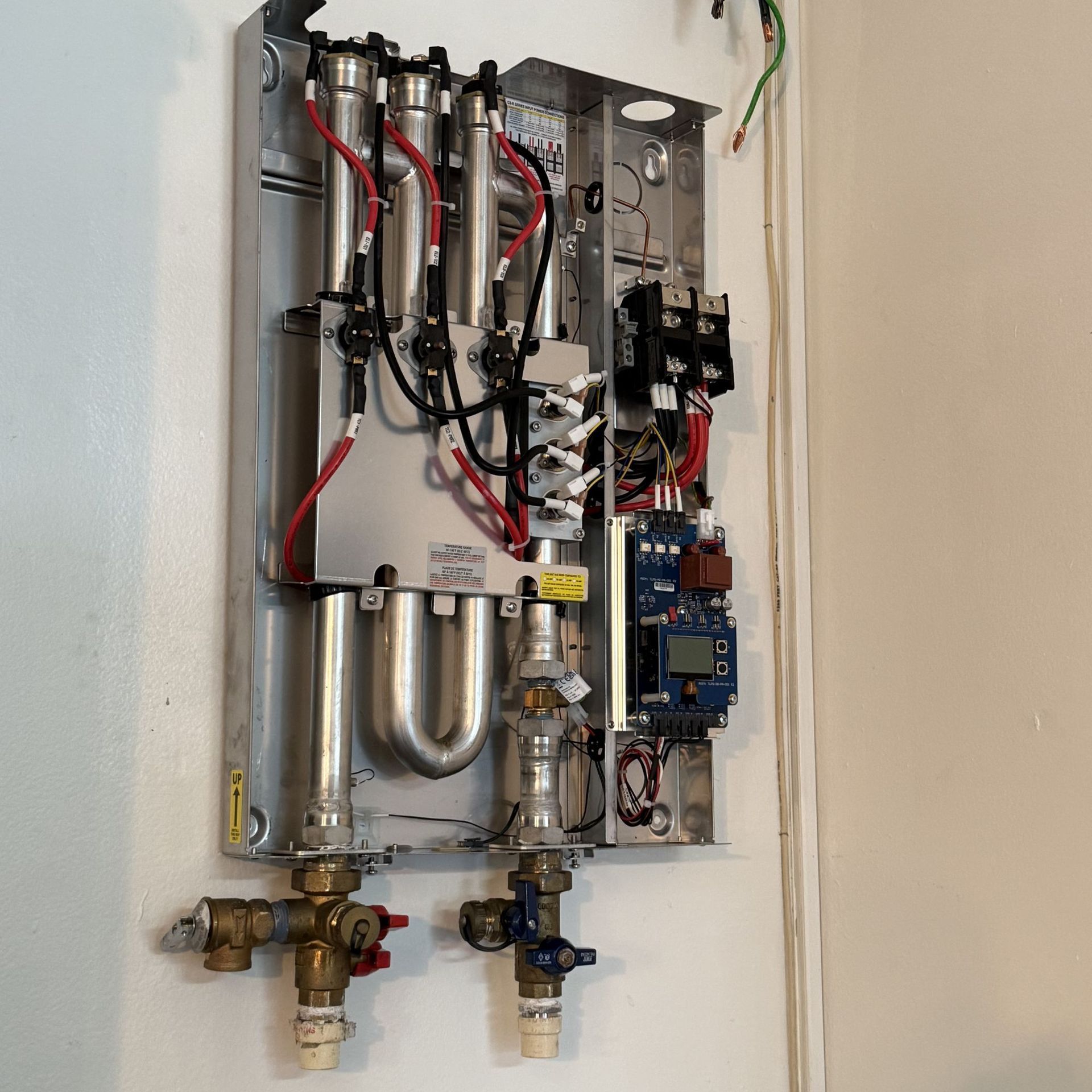 Tankless water heater, mounted on a wall, with visible pipes, wires, and electronic components.