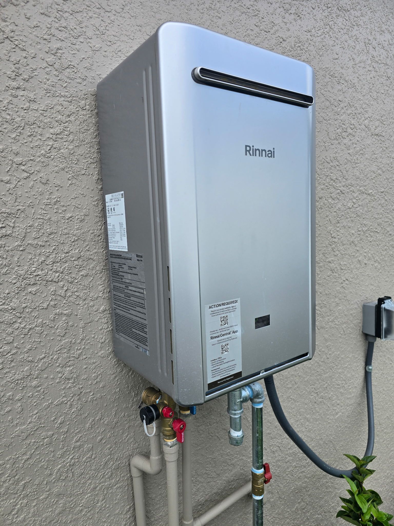 Silver Rinnai tankless water heater mounted on a stucco wall with connected pipes and electrical outlet.