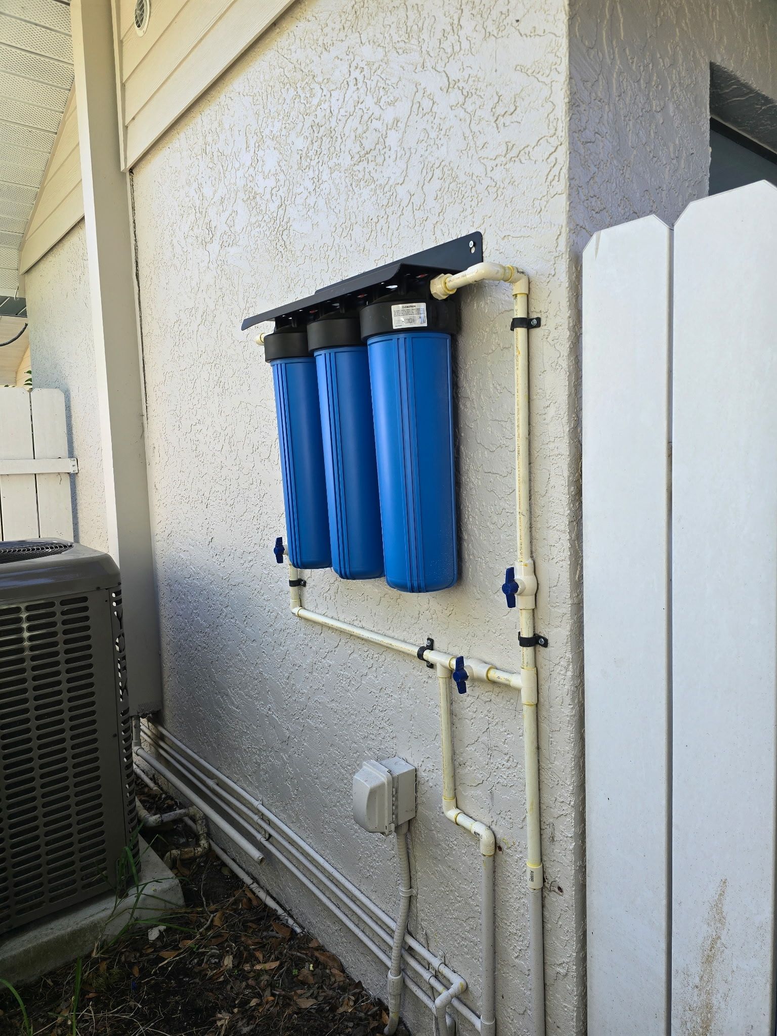 Blue water filter system installed on an exterior stucco wall with PVC piping and valves.