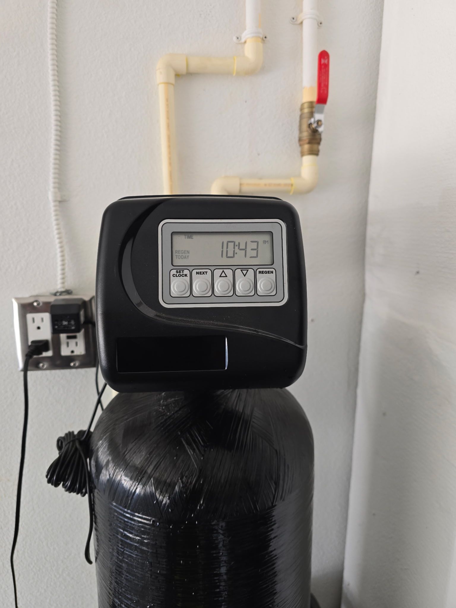 Black water softener with digital display, connected to pipes near a wall.