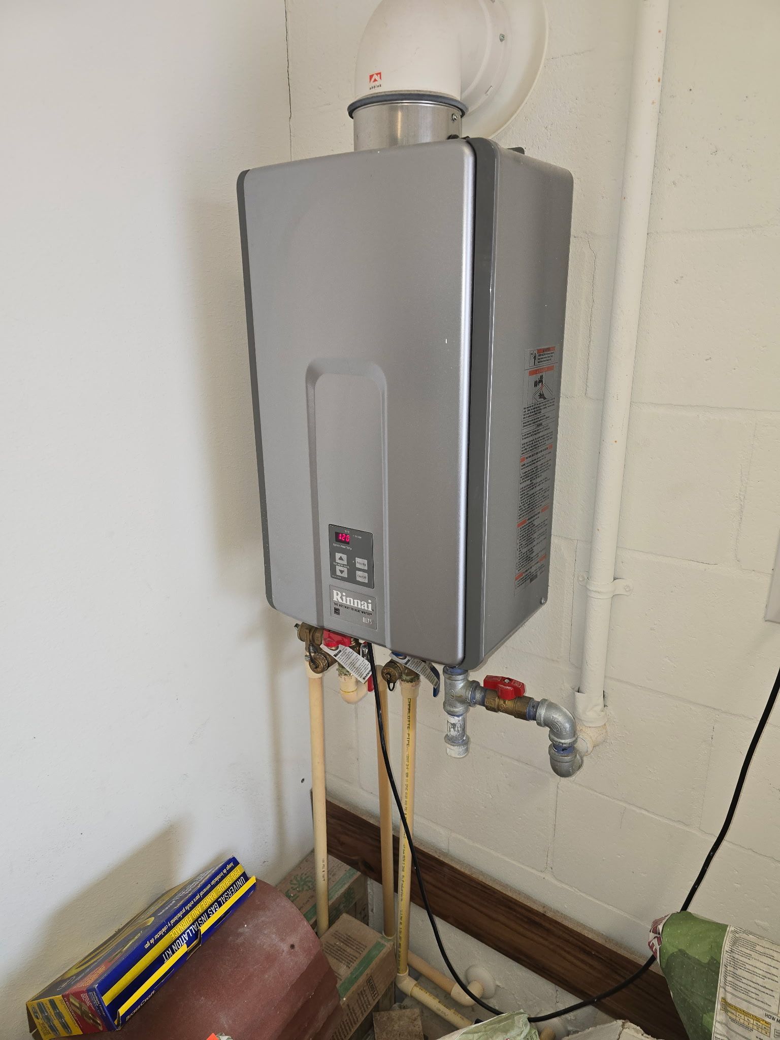 Wall-mounted gray tankless water heater with vent pipe, installed in a white-walled room with pipes.