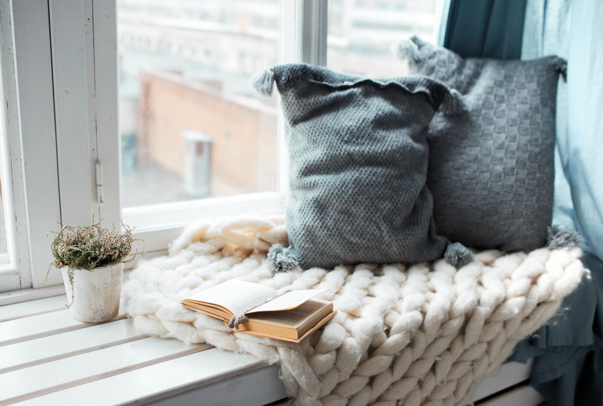 A book is sitting on a knitted blanket on a window sill.