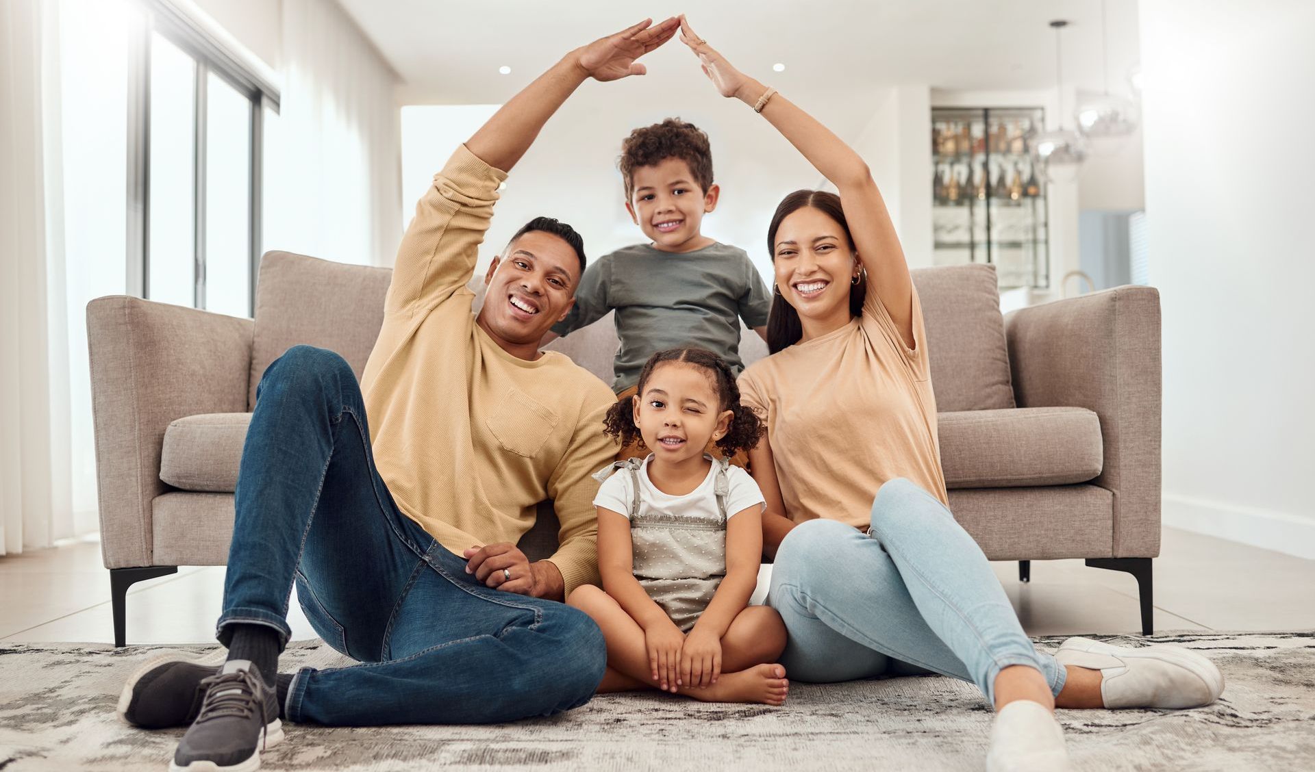 A family is sitting on the floor in a living room making a house with their hands.