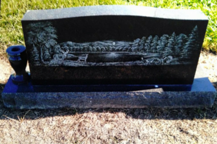 Black granite headstone featuring an etched landscape of a lake, forest, mountains, and wildlife, with a vase attached.