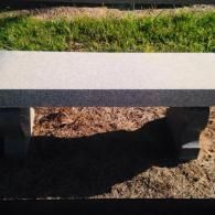 A two-toned stone bench with a light-colored seat and dark, curved legs sitting on brown mulch in an outdoor setting.