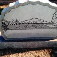 A gray stone headstone engraved with a scene of a small boat on a lake with mountains and a sunrise in the background.