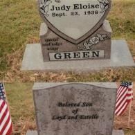 Two gravestones in a grassy cemetery. The top stone reads 
