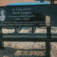 A black memorial bench for Beth Cooper, featuring her portrait and the Bible verse Matthew 19:14, set on a gravel surface.