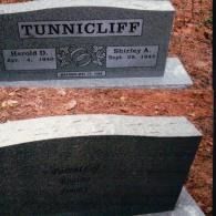 Two grey stone grave markers in a dirt cemetery setting. The top marker reads 