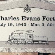 A granite gravestone for Charles Evans Fort, featuring a Masonic emblem above his dates and an engraving of a steam train.