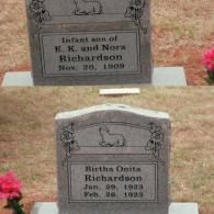 Two grey stone headstones in a cemetery, each with a carved lamb and rose motifs, situated on grass with pink flowers.