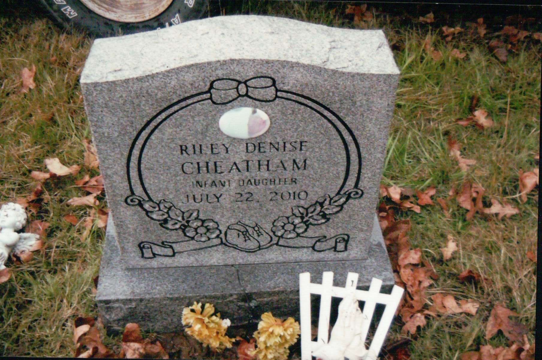 A gravestone for Riley Denise Cheatham, dated July 22, 2010, surrounded by grass, fallen leaves, and small decorations.