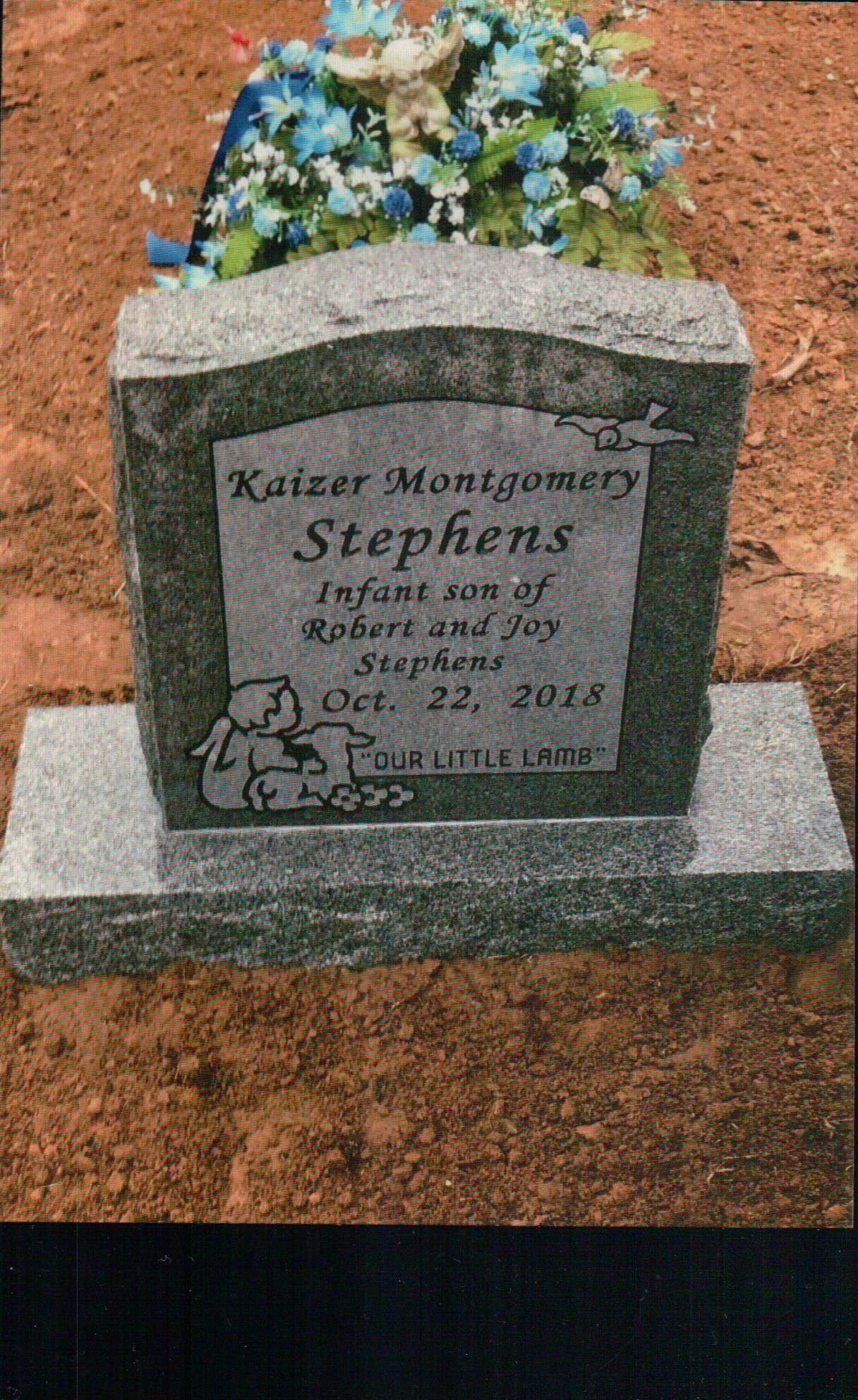 A granite headstone for infant Katzer Montgomery Stephens, dated Oct. 22, 2018, with floral tributes on red soil.