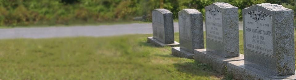 A row of four grey granite tombstones stands in a grassy field next to a paved road on a sunny day.
