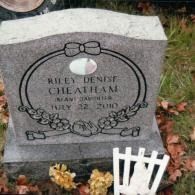 A granite headstone for Riley Denise Cheatham, dated July 22, 2010, with floral and ribbon engravings, in a cemetery.