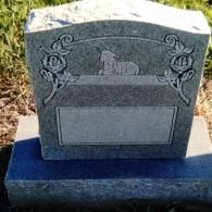 A grey granite headstone featuring carved floral designs on the sides and a relief of a resting lamb in the center.