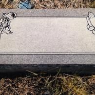 A grey granite grave marker with a rose engraving on the left, praying hands on the right, and a blank rectangular center.