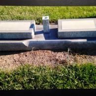 A stone cemetery marker with two raised rectangular panels and a central flower vase holder, surrounded by grass.