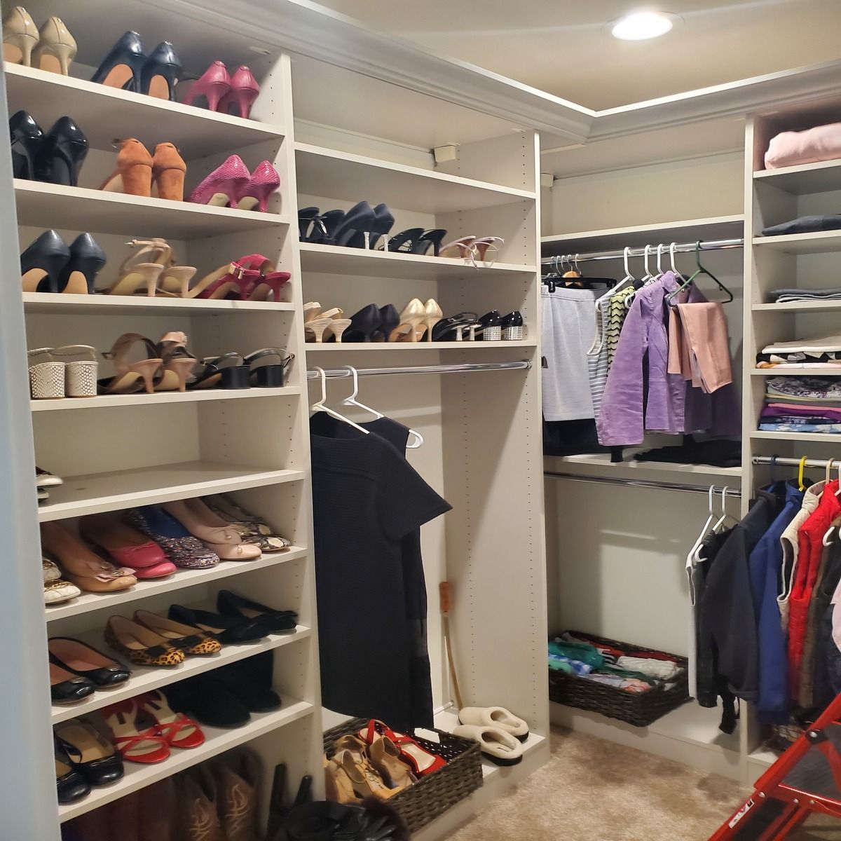 Walk-in closet with shelves full of shoes, hanging clothes, and a basket of items. White shelving, neutral carpet, and various clothing items.