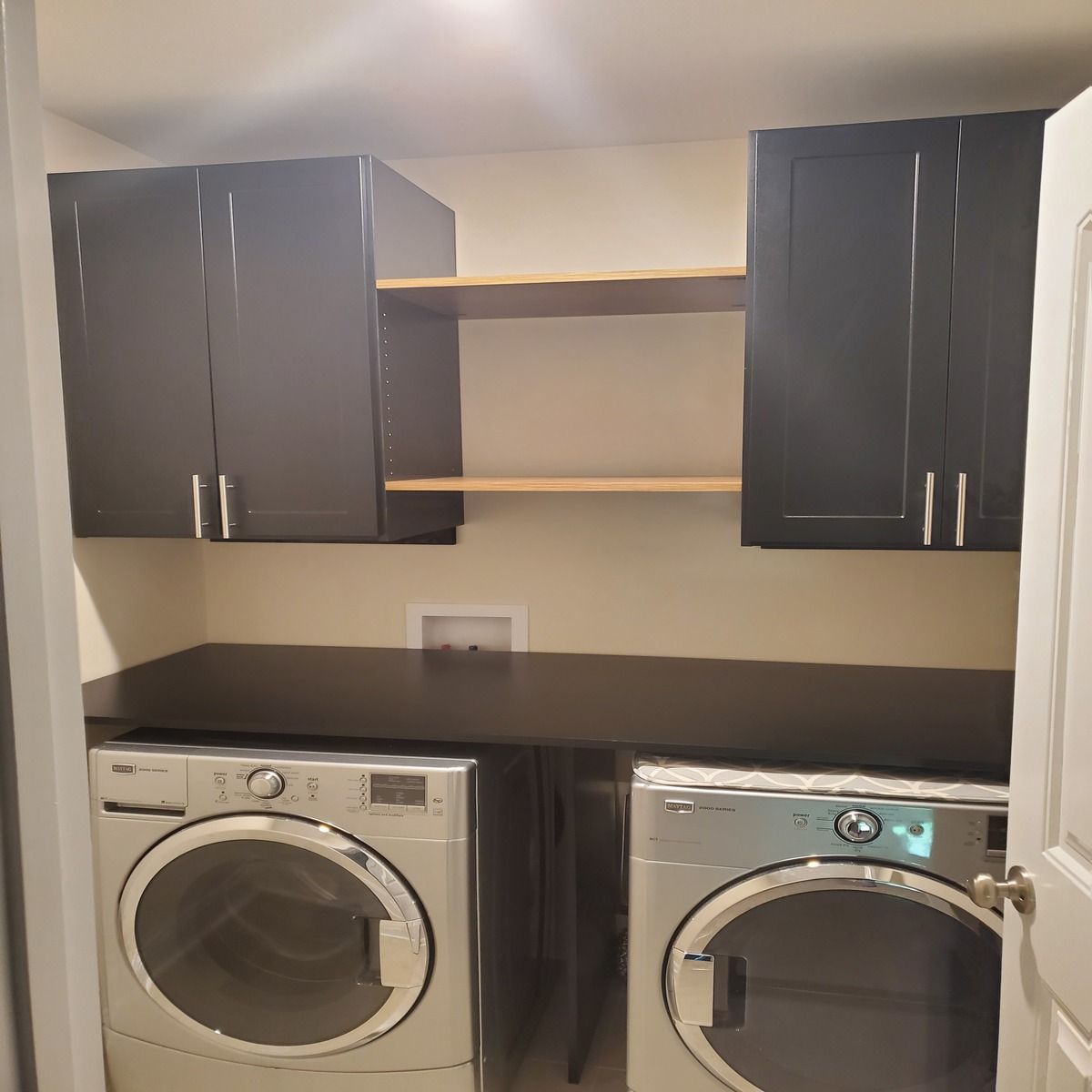 A laundry room with a washer and dryer below dark cabinets and shelves. The room has a dark countertop and a partially visible doorway.