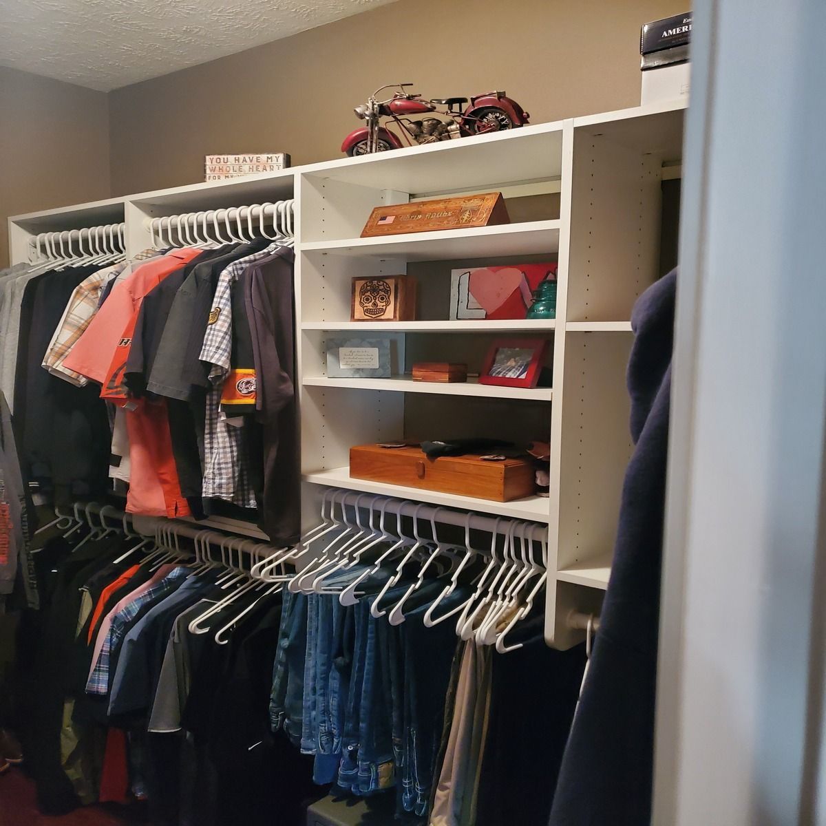 A walk-in closet with white shelves and hanging clothes. Shelves hold decorative items, and a motorcycle model sits on top.