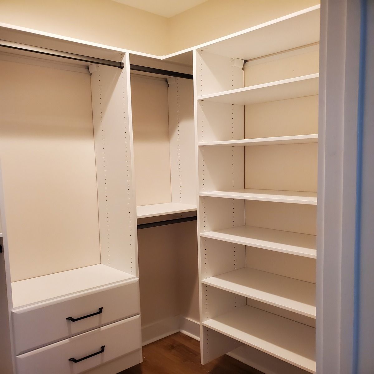Empty, white walk-in closet with shelves, drawers, and a hanging rod. Corner shelving unit is on the right.