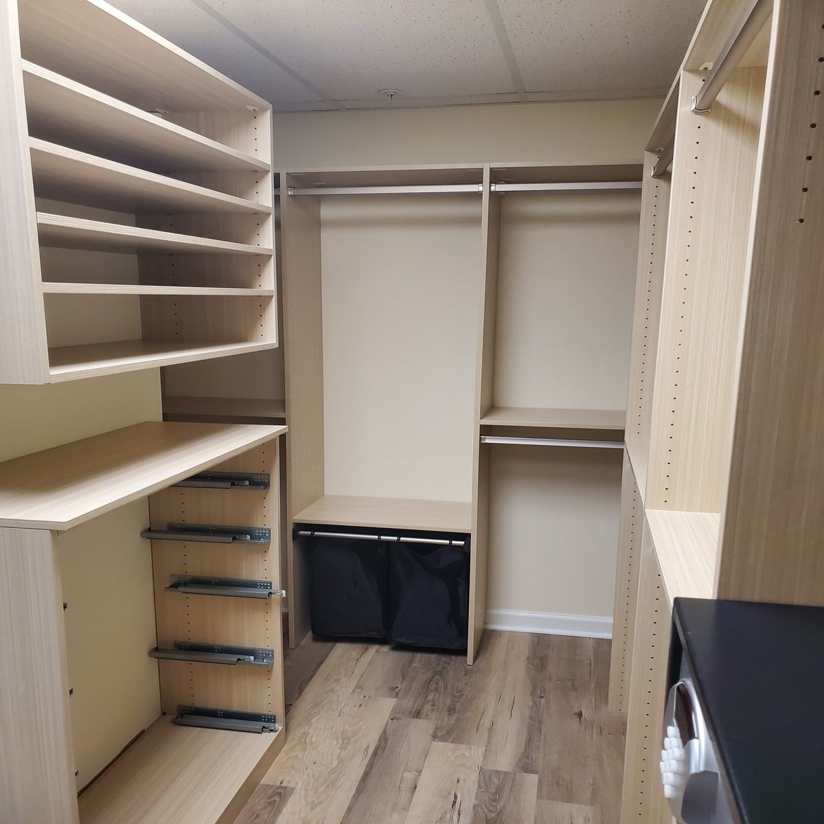 A well-organized walk-in closet with light wood shelving, drawers, and hanging rods. The closet features a black hamper and wooden floors.