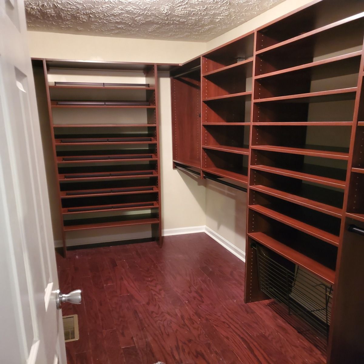 Empty walk-in closet with dark wood shelves and flooring. Features include a door, wall-mounted shelves, and wire basket.