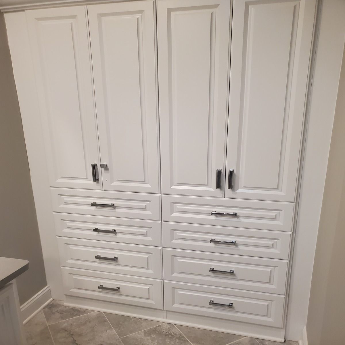 White built-in cabinet with four upper doors and four sets of drawers. The cabinet has black handles and is set in a light-colored room with stone-like flooring.