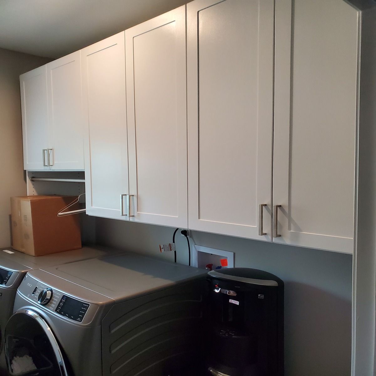 White laundry room cabinets above a washing machine and dryer with a coffee maker, power outlets, and a brown cardboard box.