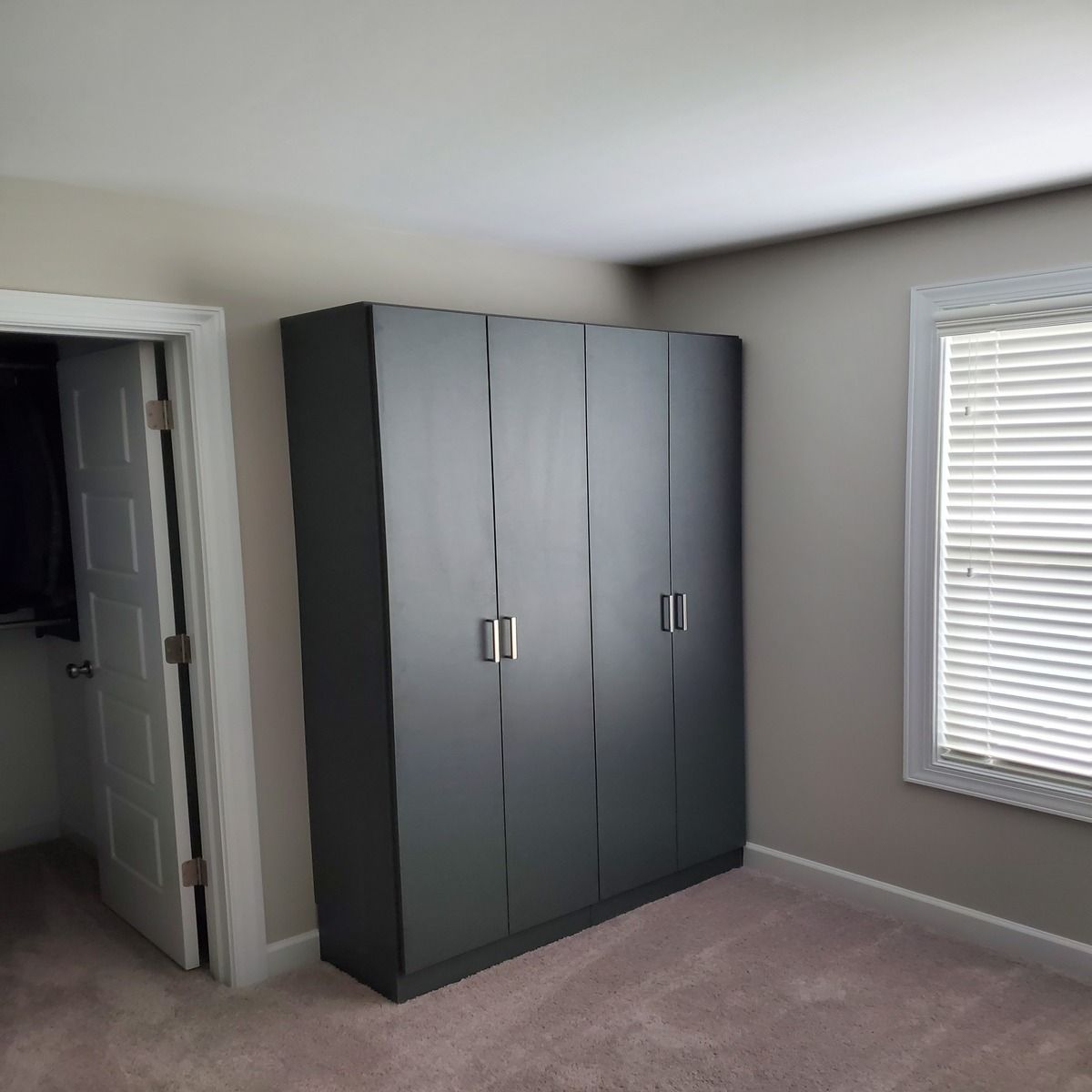 A dark gray wardrobe with four doors stands in a bedroom next to a door and a window with blinds.