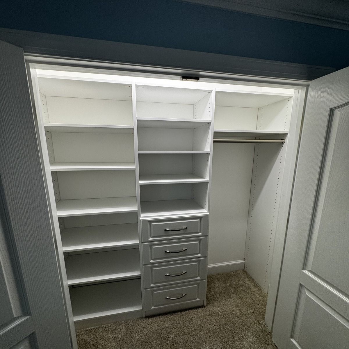 Open white closet with built-in shelves, drawers, and a rod for hanging clothes. Carpeted floor.
