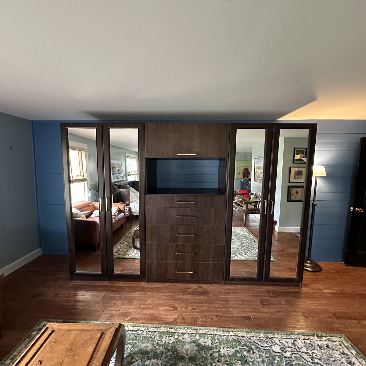 Dark wood built-in wardrobe with mirrored doors and a center section for a TV and drawers. The setting is a room with wood floors and blue accent walls.
