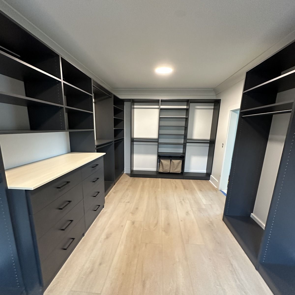 A spacious walk-in closet with dark shelving, drawers, and light wooden flooring. Includes hanging rods, shelves, and storage baskets.