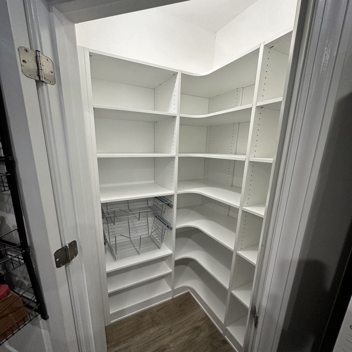 White pantry with corner shelves, open door, wire basket, and wooden floor.