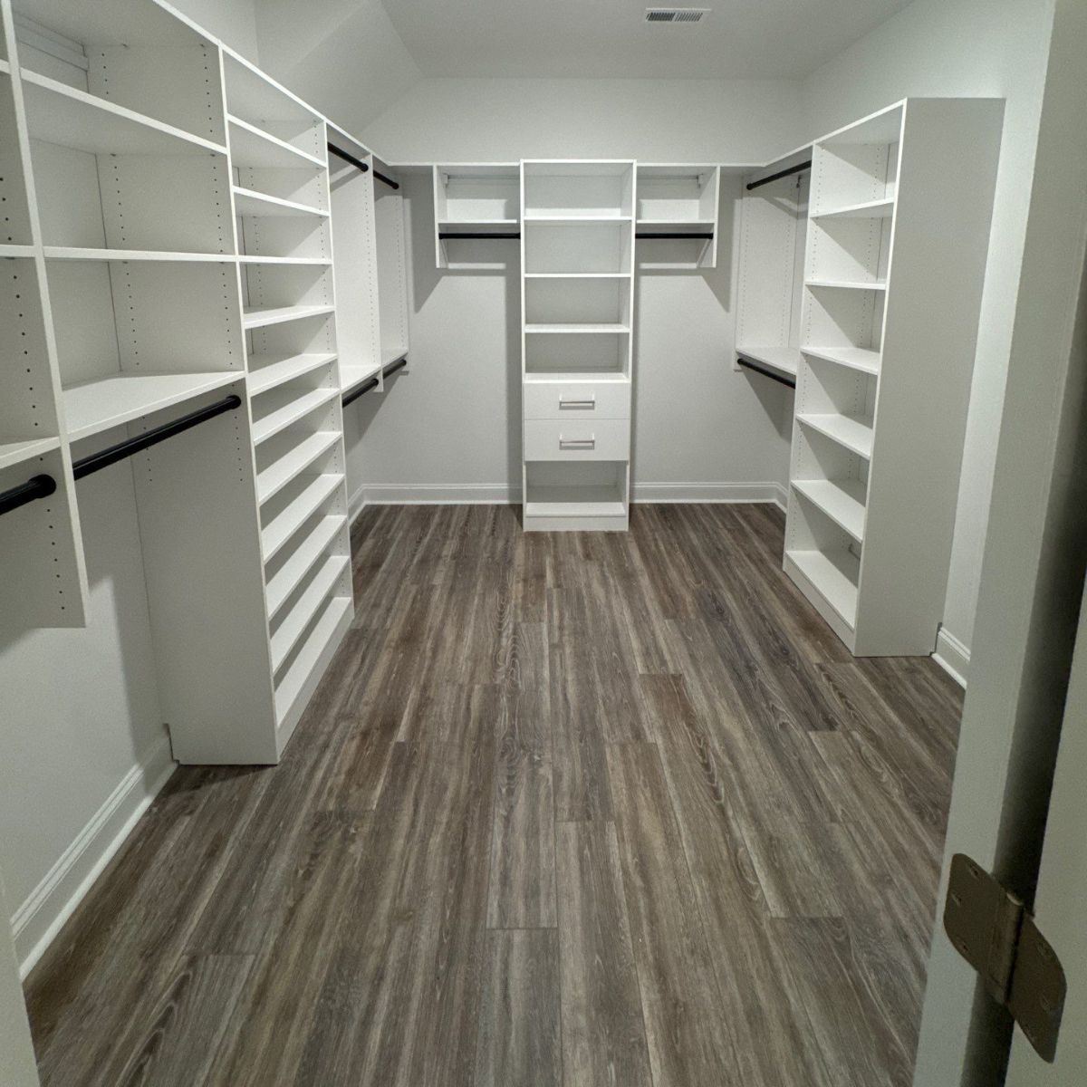 Empty walk-in closet with white shelving, hanging rods, and wood-look floor.