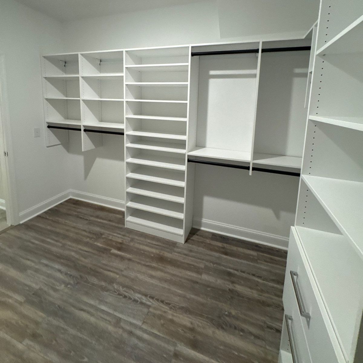 Empty white walk-in closet with wooden shelves and flooring. A pair of drawers are visible on the right.