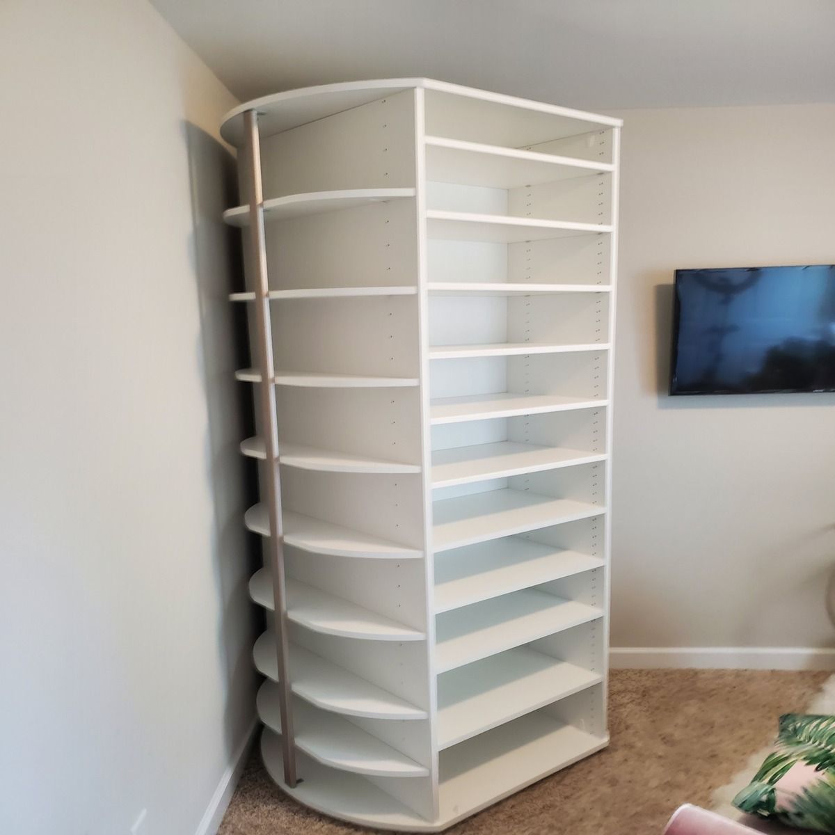 White corner shelving unit with curved sides and multiple shelves against a beige wall. A TV is visible on the wall nearby.